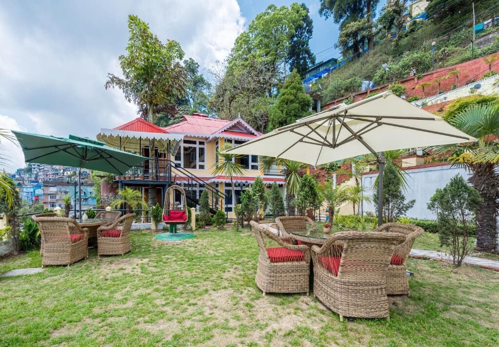 Un patio con muebles de mimbre y sombrillas, rodeado de abundante vegetación y un edificio de techo rojo.