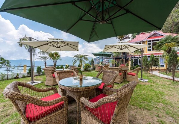 An outdoor seating area with wicker furniture and green umbrellas, overlooking a body of water and mountains. - Udaan Nirvana Resort Darjeeling (Darjeeling)