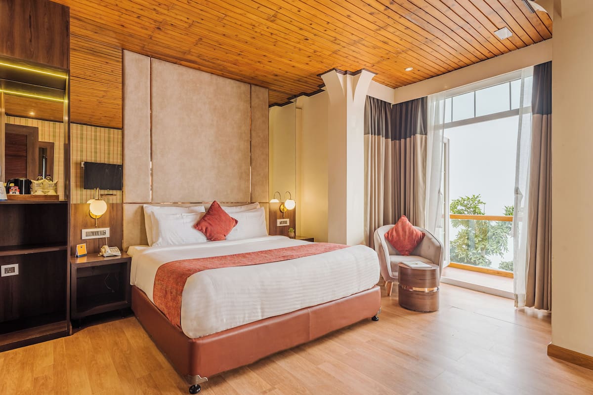 Een hotelkamer met een groot bed, houten plafond, een televisie, bedlampen, een stoel en een balkon met gordijnen.