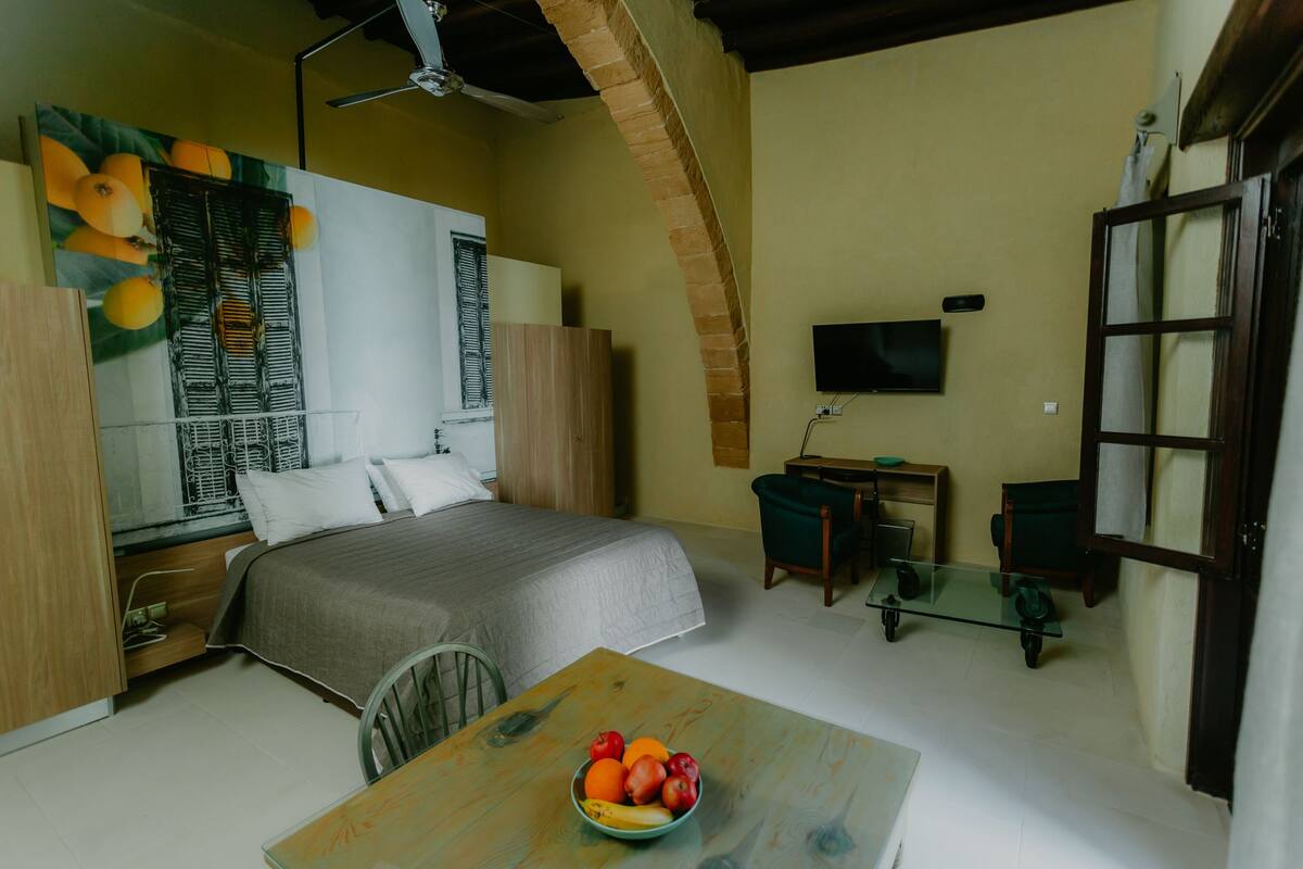 A bedroom with a bed, a desk, a TV, and a dining table with fruit.