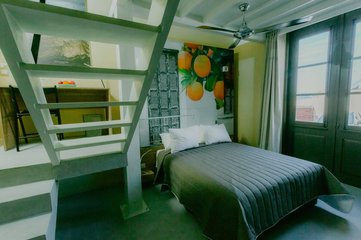 A bedroom with a bunk bed, a bed with a dark-colored bedspread, a ceiling fan, a window with curtains, and a wall with a fruit-themed mural.