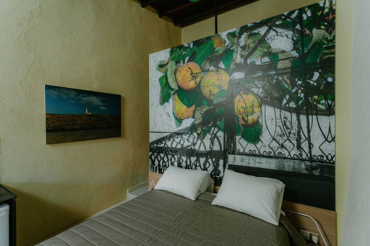 A bedroom with a bed, pillows, a painting of fruit, and a smaller picture on the wall.