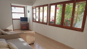 Ein Wohnzimmer mit einer Couch, einem Fernseher und einem Fenster mit Blick auf Grünflächen.