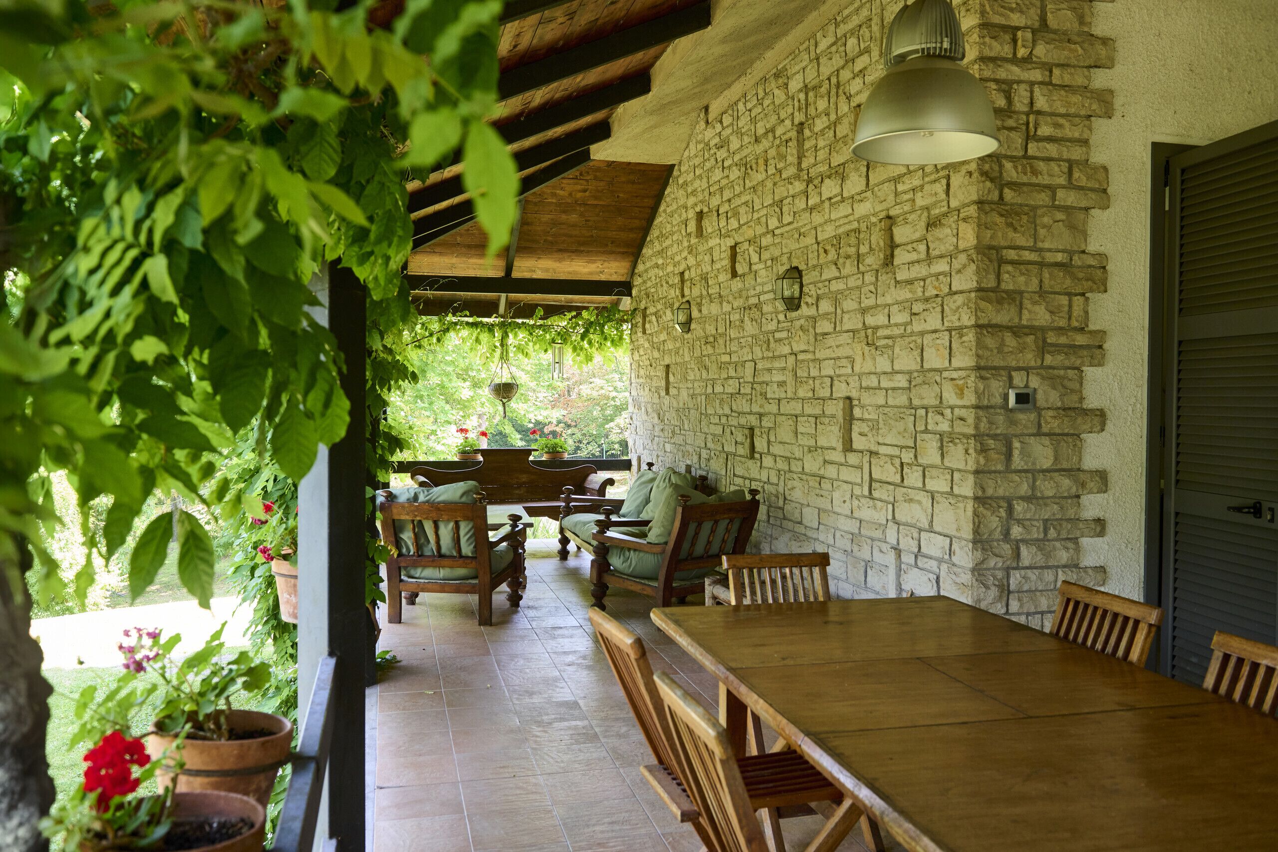 Une terrasse couverte avec un mur en pierre, des meubles en bois et des plantes suspendues.