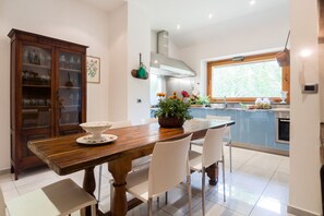 A dining area with a wooden table, white chairs, a cabinet with glass doors, and a kitchen with blue cabinets and a window.