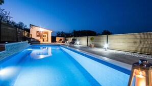 A swimming pool with a wooden deck, lounge chairs, and a building with illuminated windows. - Cozy family-friendly fenced villa, pool and outdoor jacuzzi (Karojba)