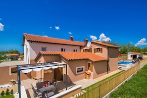 Uma casa de dois andares com telhado em telhas, um pátio com mesa e cadeiras, e uma piscina.