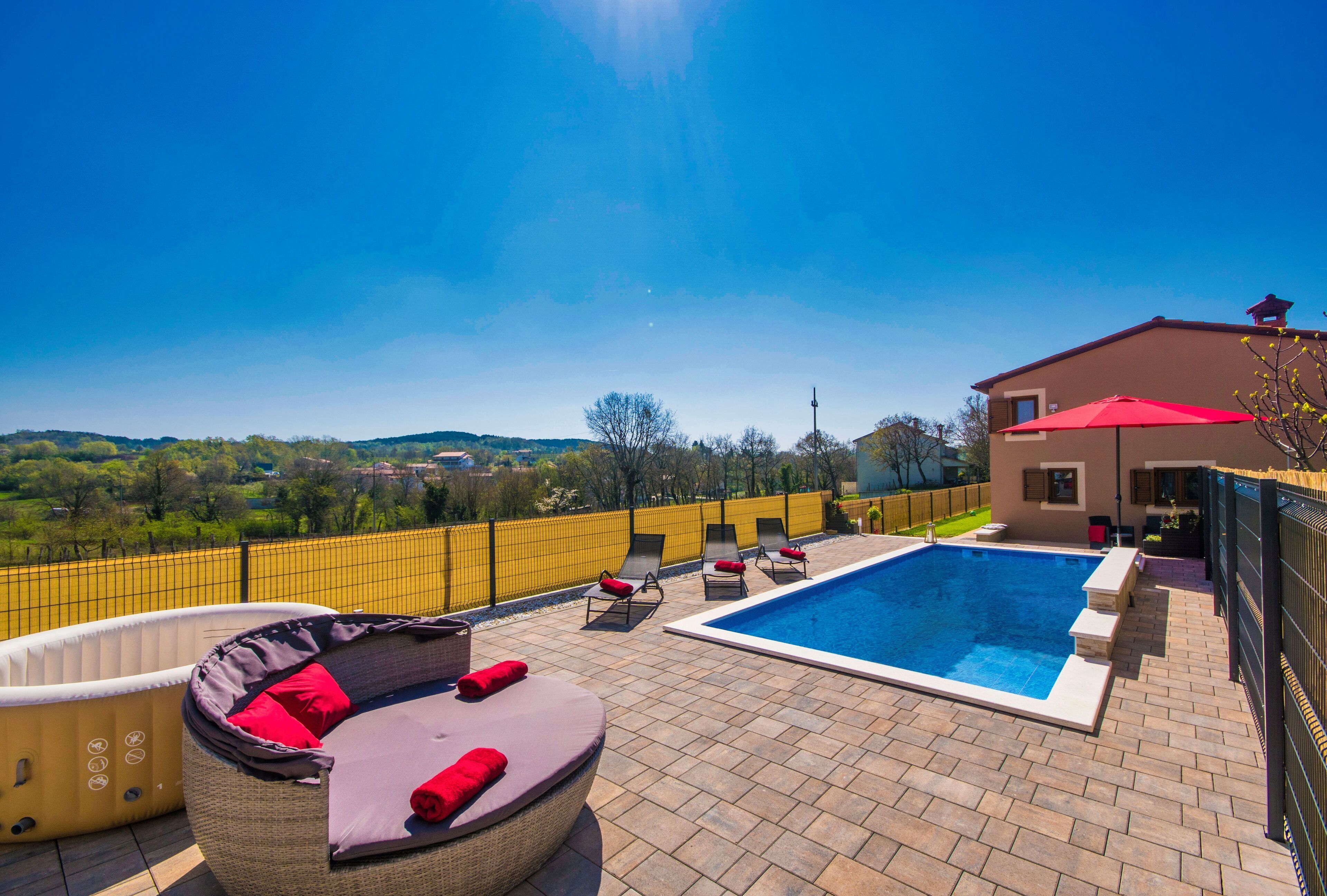 A rooftop swimming pool with lounge chairs and a hot tub, surrounded by a yellow fence and a view of the countryside.