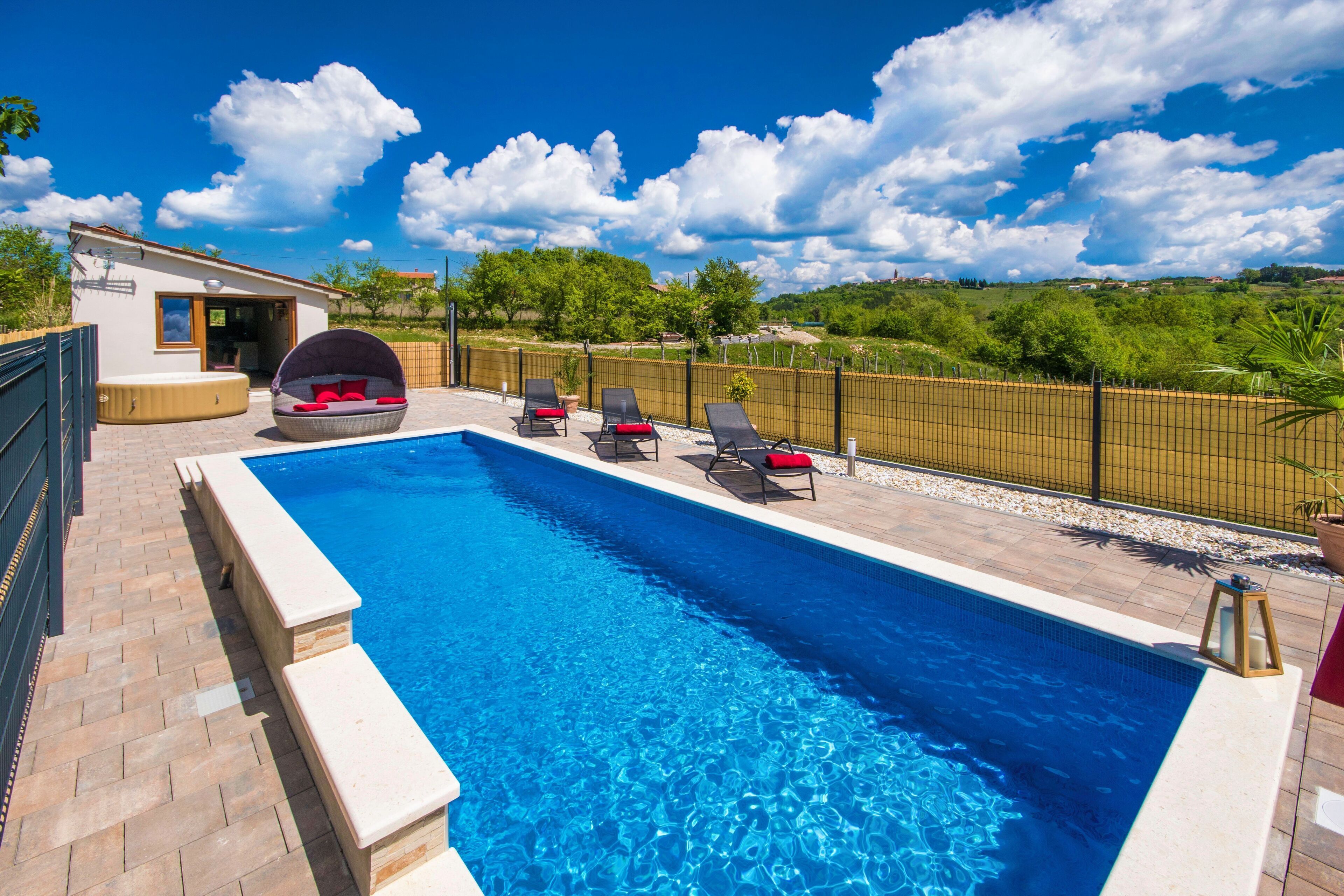 A swimming pool with lounge chairs and a hot tub, surrounded by a fence and greenery.