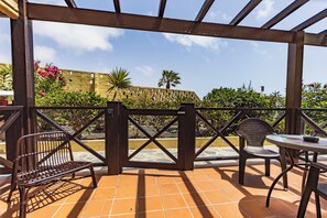 Una veranda esterna con una pergola in legno, due sedie in metallo, un tavolo rotondo e vista su un edificio giallo e del verde.
