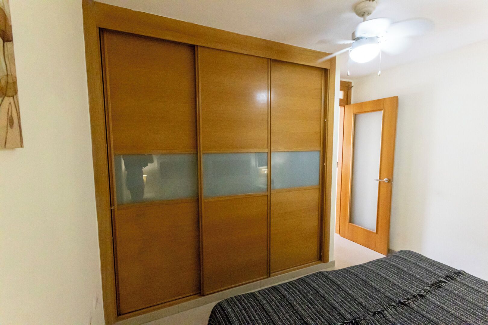 Un dormitorio con un armario de madera, una cama con manta gris y un ventilador de techo.