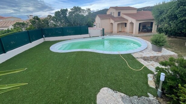 Un jardin avec une piscine, une pelouse artificielle verte, un chemin de pierres et une maison avec un toit en tuiles.
