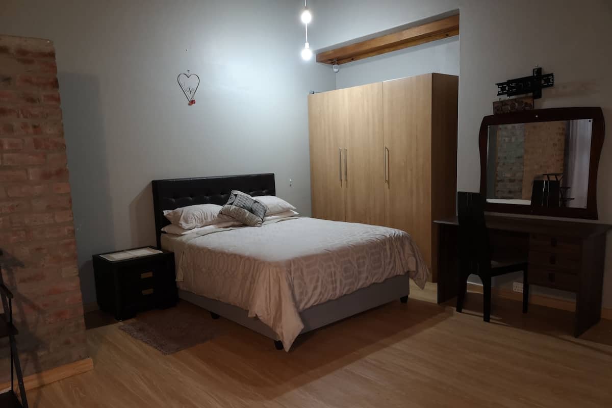 A bedroom with a bed, nightstands, a wooden wardrobe, a desk with a mirror, and a brick wall.