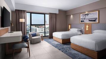 Quarto de hotel com cama, escrivaninha, poltrona, televisão e uma grande janela com vista para a cidade.