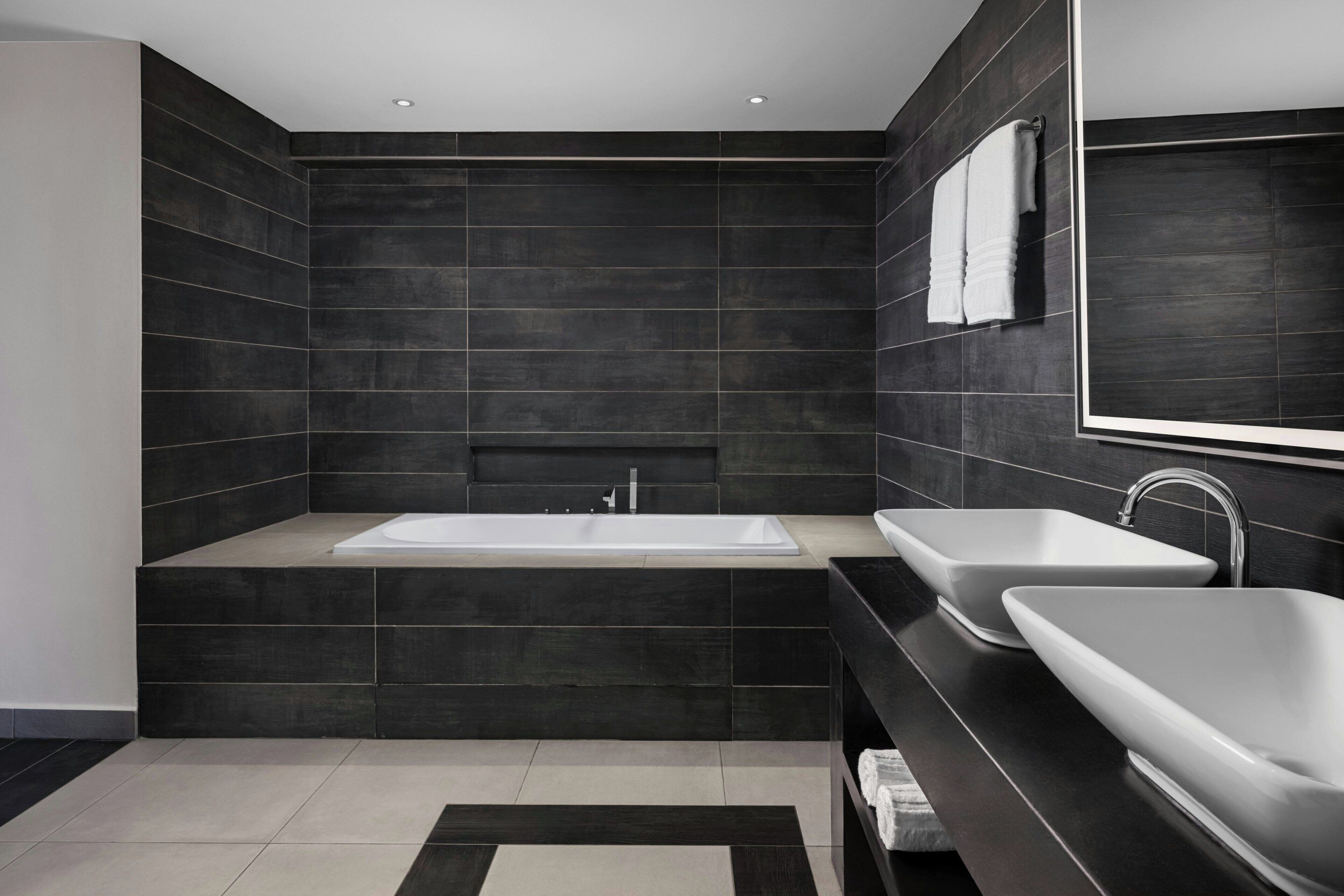 A modern bathroom with dark tiled walls, a large bathtub, and two sinks.