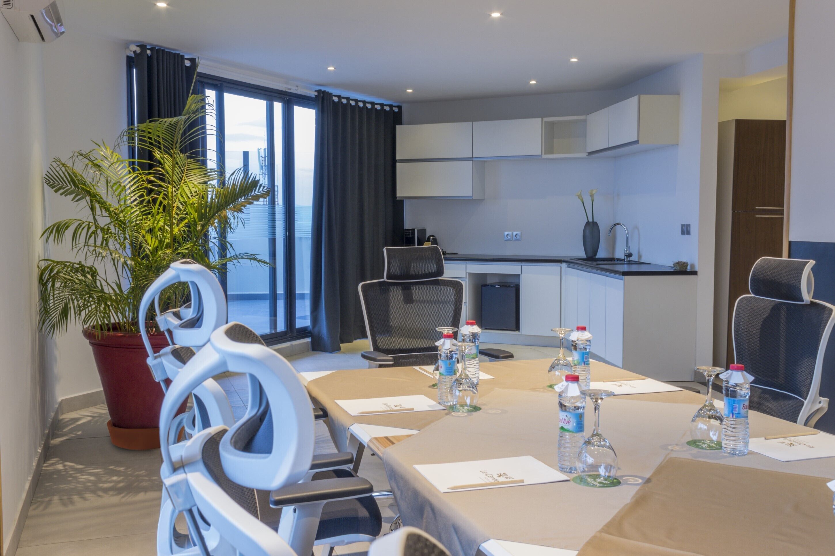 Habitación de hotel moderna con una mesa de comedor preparada para seis personas, equipada con botellas de agua y vasos, una planta en maceta y vistas de la ciudad a través de grandes ventanales.