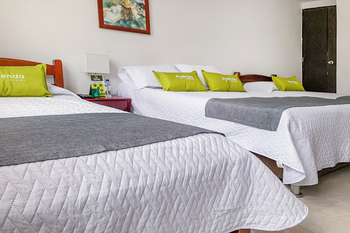 A hotel room with two beds, each with white bedding and green pillows, a wooden headboard, a nightstand with a lamp, and a painting on the wall.