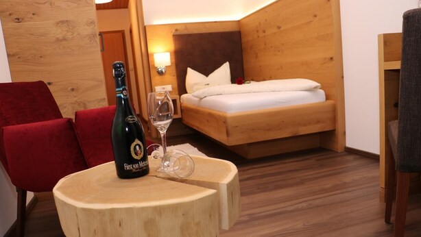 A cozy room with a wooden bed, a red chair, a small wooden table with a bottle and glass, and a fluffy rug. - Gasthof-Pension Kerber (Fürstenstein)