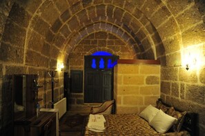 A stone-walled room with a bed, a chair, a desk, and a door with a blue-lit window. - Osmanoglu Hotel (Guzelyurt)