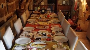 Una mesa de comedor con platos, vasos y diferentes alimentos, rodeada de sillas en una habitación rústica con paredes de piedra.