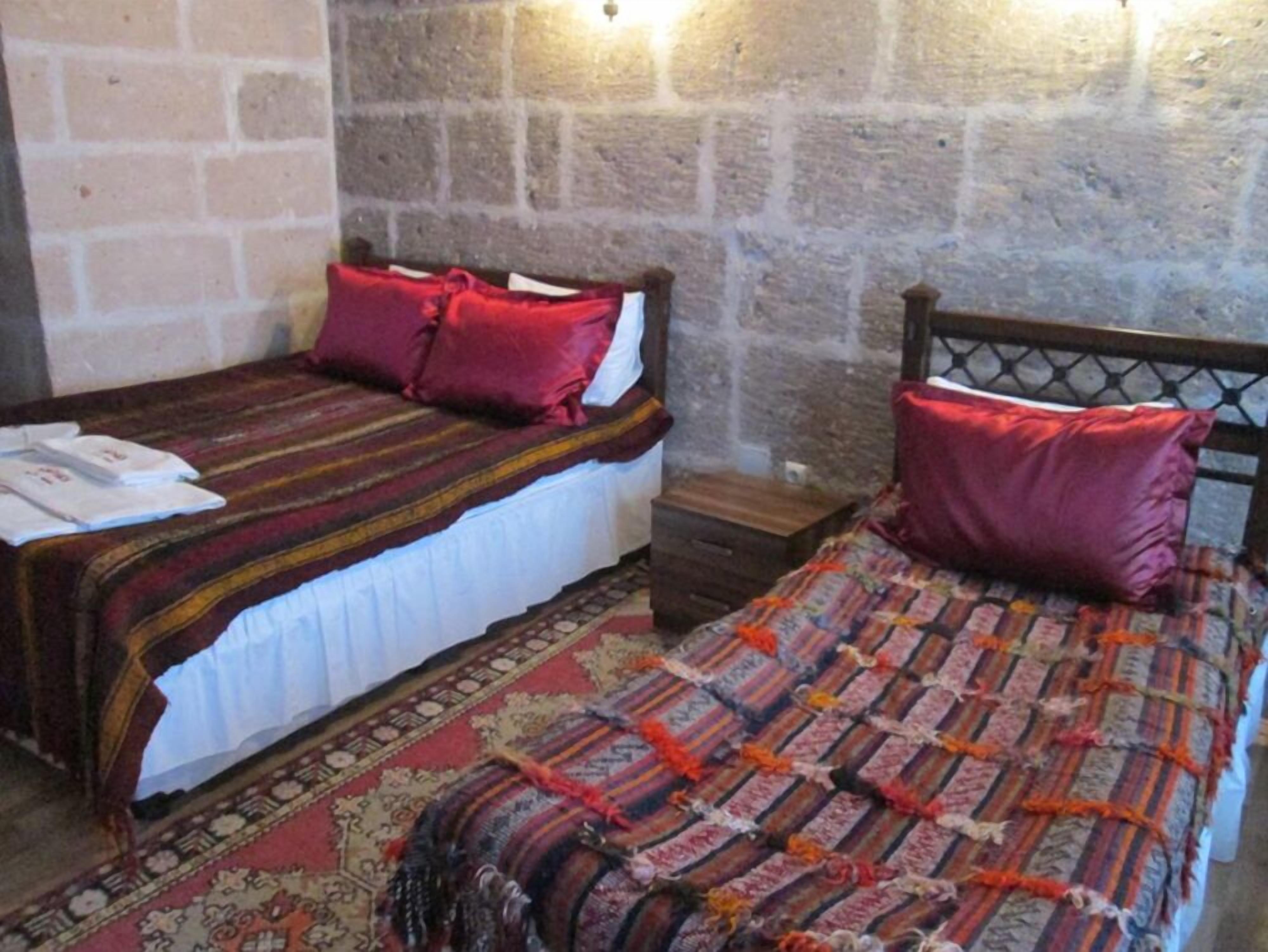 Une chambre avec deux lits, chacun recouvert d’une couvre-lit et agrémenté d’oreillers aux motifs différents. On y trouve des tables de chevet en bois et une petite table en bois. Les murs sont faits de blocs de pierre.