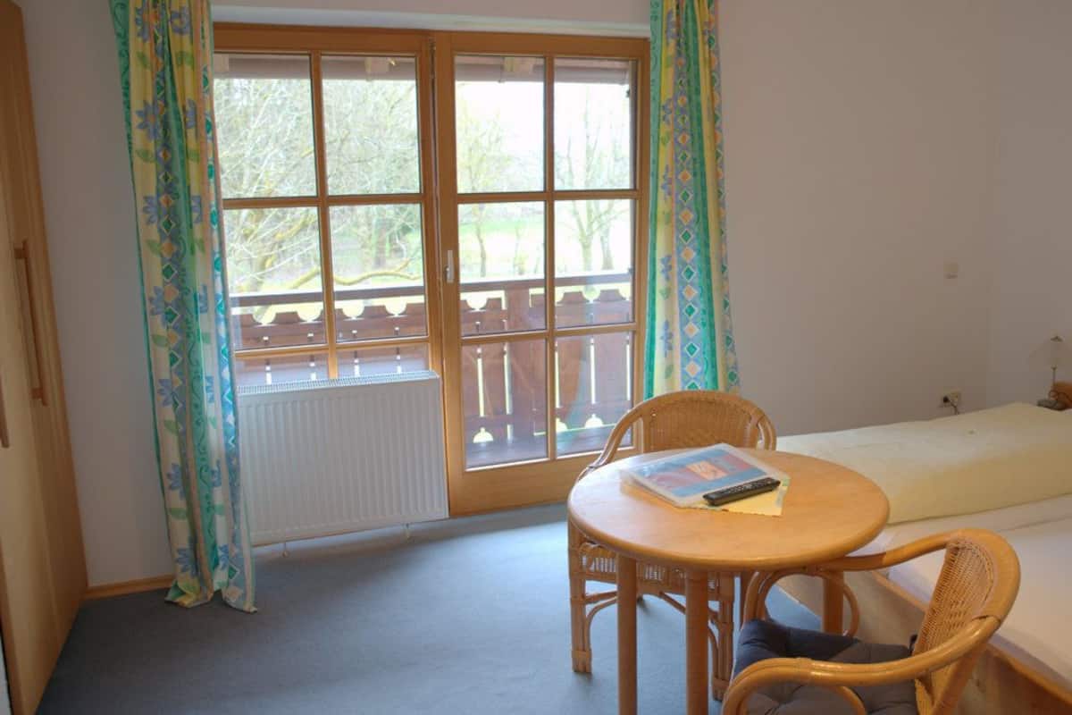 A room with a bed, a small table with a book, a chair, and a window with curtains.