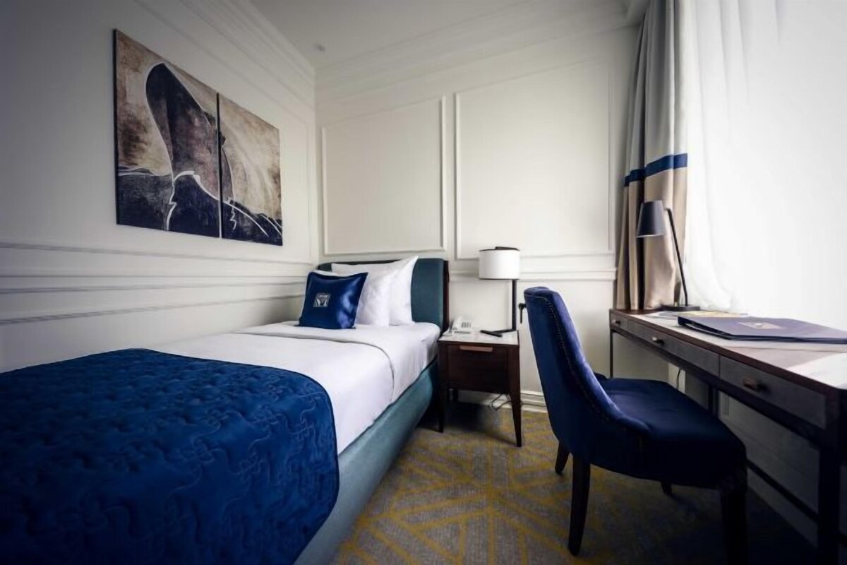 A hotel room with a bed, a desk, a chair, a lamp, and artwork on the wall.