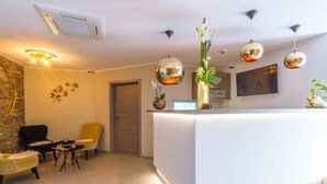 A modern reception area with a white reception desk, black chairs, a small round table, and decorative wall art. - ManavaHotel (Herstal)