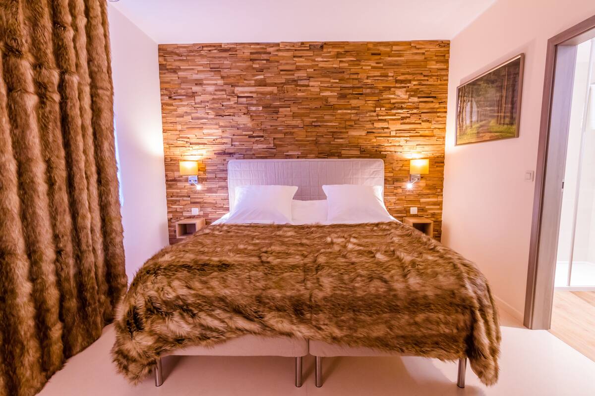 Un dormitorio con cabecera de pared de piedra, colcha marrón y mullida, y dos lámparas fijadas a la pared.