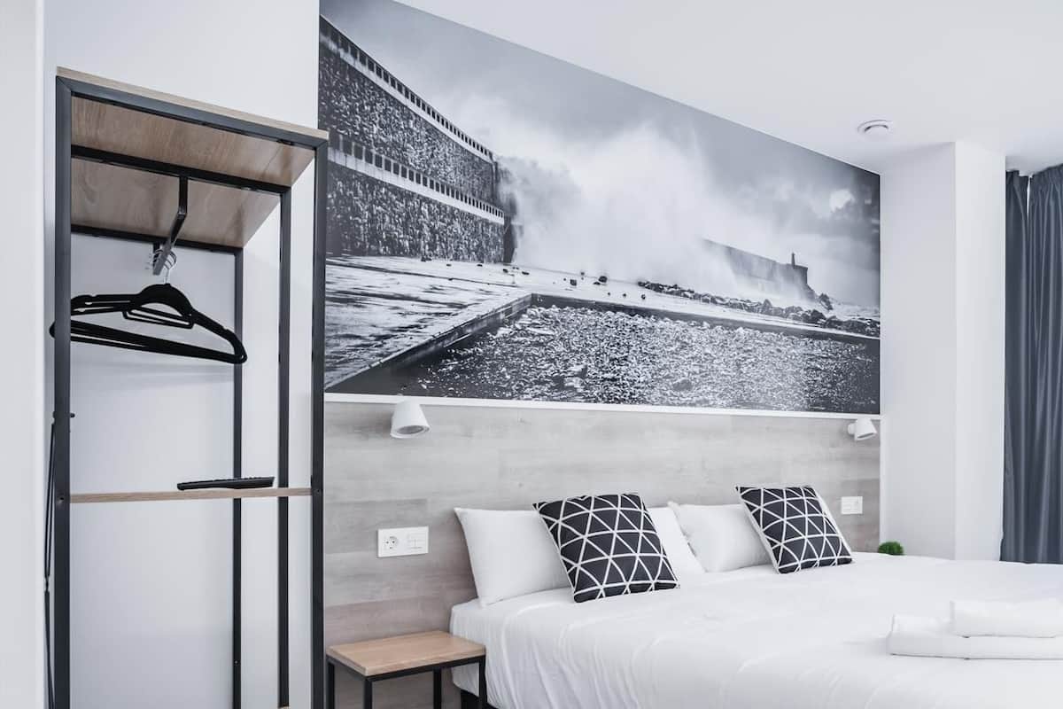 A modern hotel room with a large bed, a wardrobe, a small table, and a wall-mounted shelf.