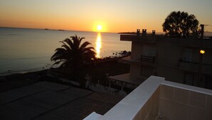 Vista al atardecer desde un balcón con vista a un mar tranquilo, con una palmera y un edificio de tejado a la vista.