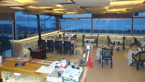 Un restaurante con mesas y sillas dispuestas para comer, amplias ventanas que ofrecen vista a la ciudad, y una barra con diferentes productos.