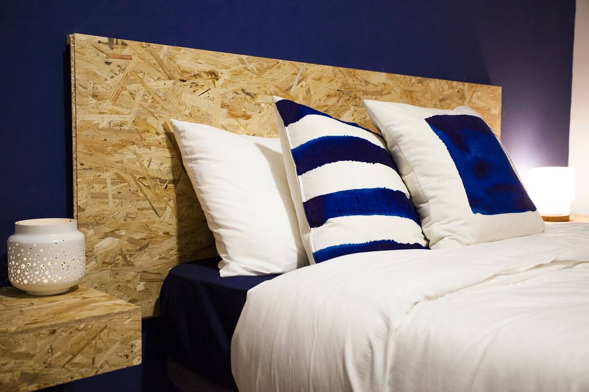 A bed with white and blue striped pillows, a wooden headboard, and a white vase on a wooden nightstand.