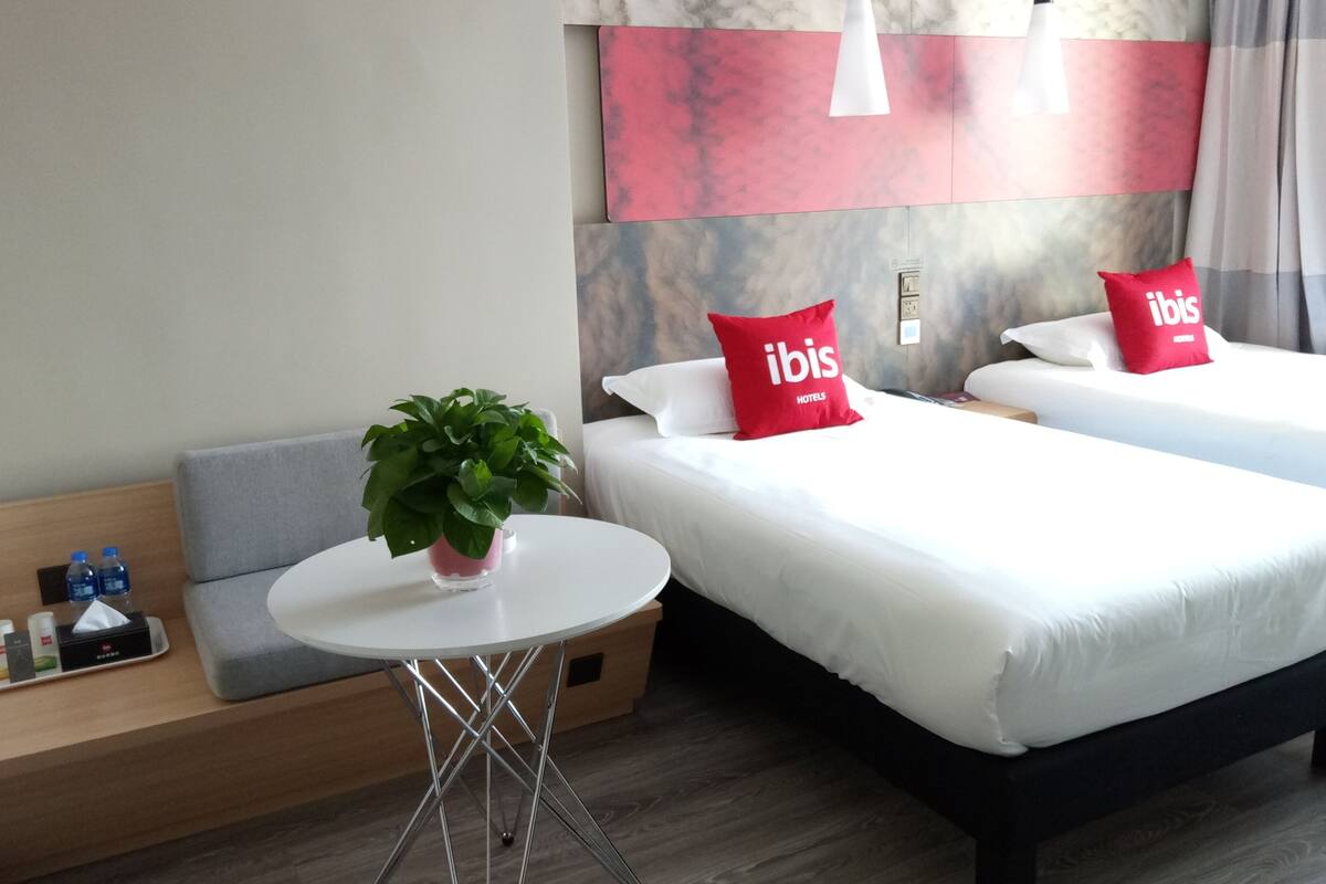 A hotel room with two beds, a small table with a plant, a bedside table with water bottles, and a grey sofa.