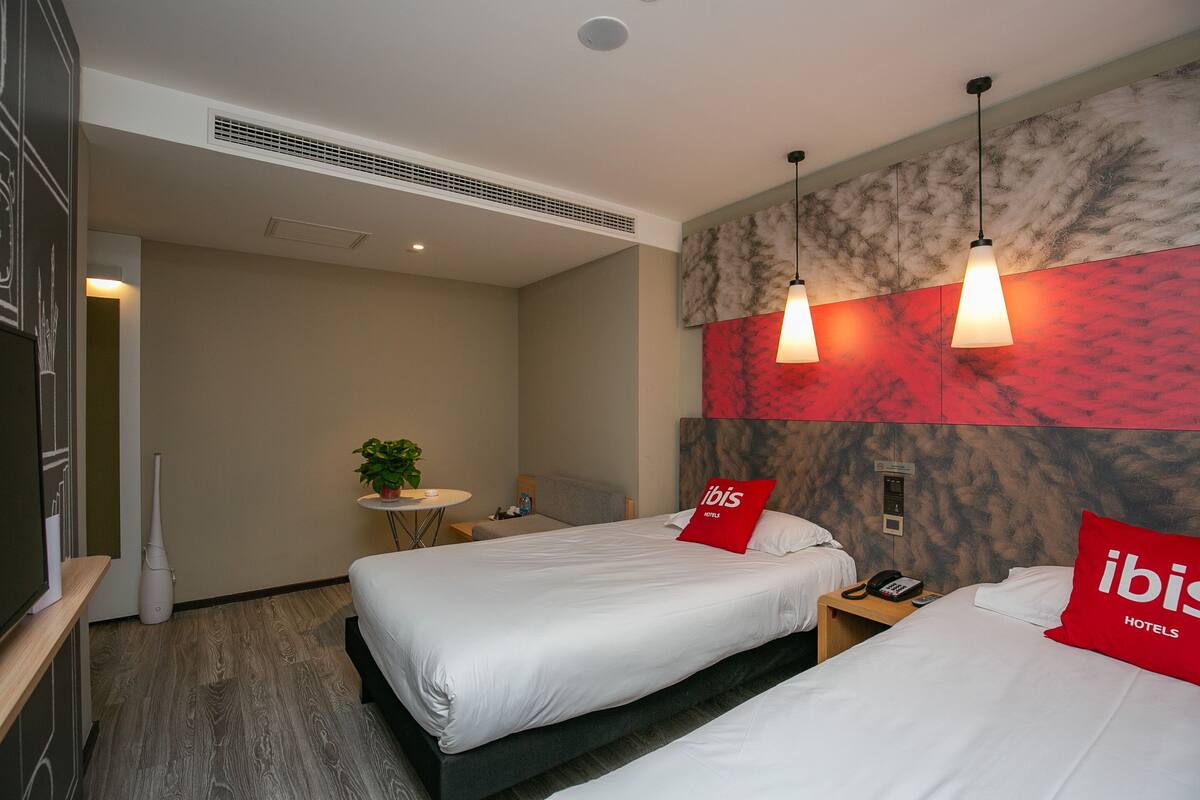 A hotel room with two beds, a small table with a plant, a wall-mounted heater, and a red pillow with the hotel logo.