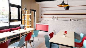A modern cafe with red and blue chairs, white tables, and a kitchen area with utensils and appliances.
