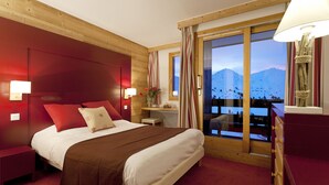 A hotel room with a large bed, wooden headboard, a nightstand, a dresser, a window with a mountain view, and a lamp.
