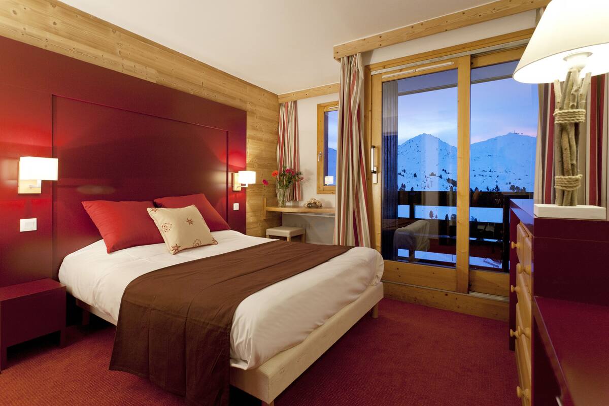 Habitación de hotel con una cama grande, cabecera de madera, una mesita de noche, un aparador, una ventana con vistas a la montaña y una lámpara.