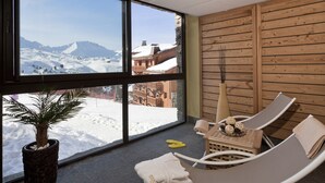 A room with large windows offering a view of snow-covered mountains, a wooden wall, a lounge chair, and a potted plant.