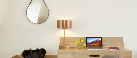 A hotel room with a wooden desk, a laptop, a lamp, a mirror, a vase with flowers, and a black bag on a shelf.
