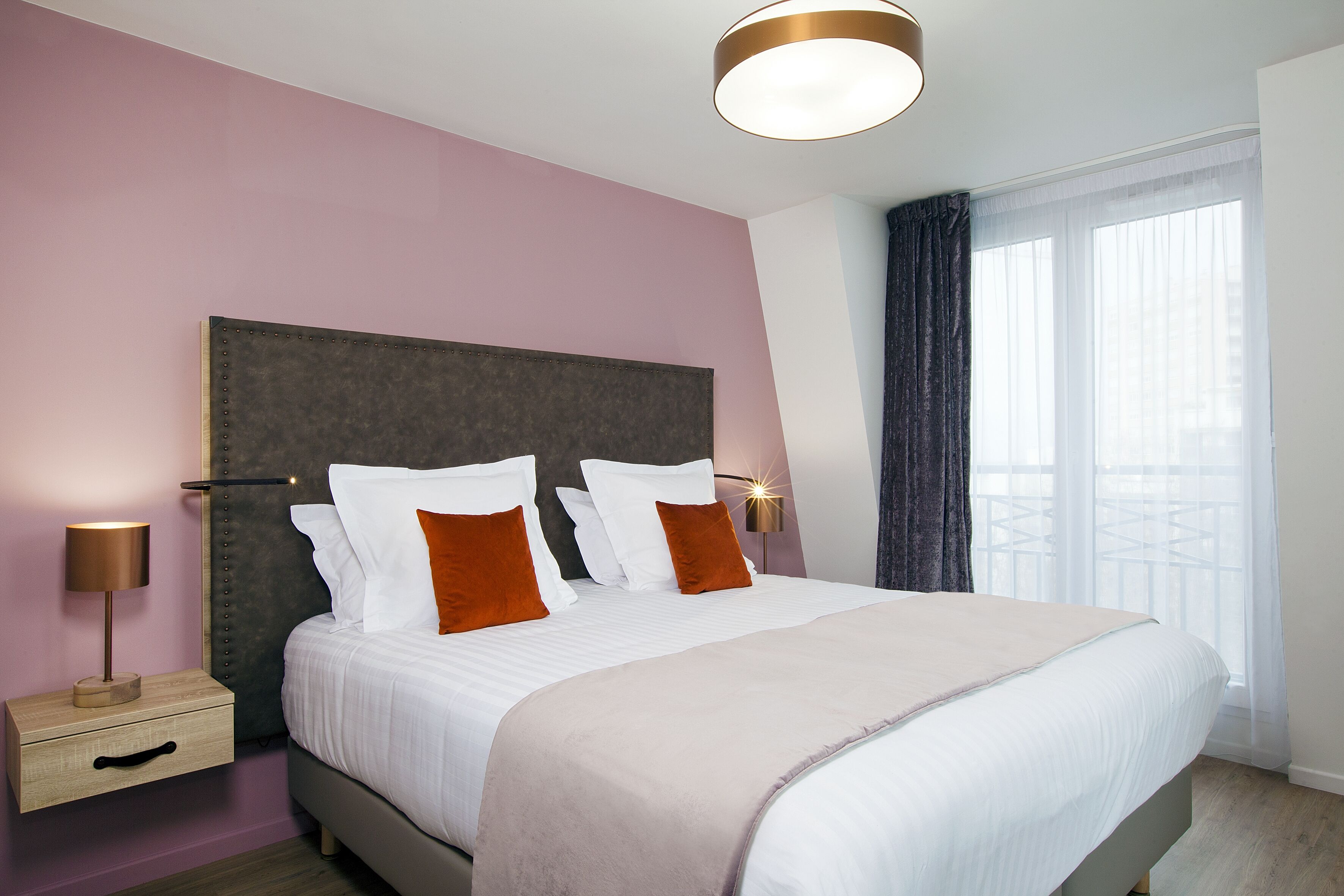 A neatly made bed with white and orange pillows, a wooden nightstand with a drawer, and a bedside lamp.
