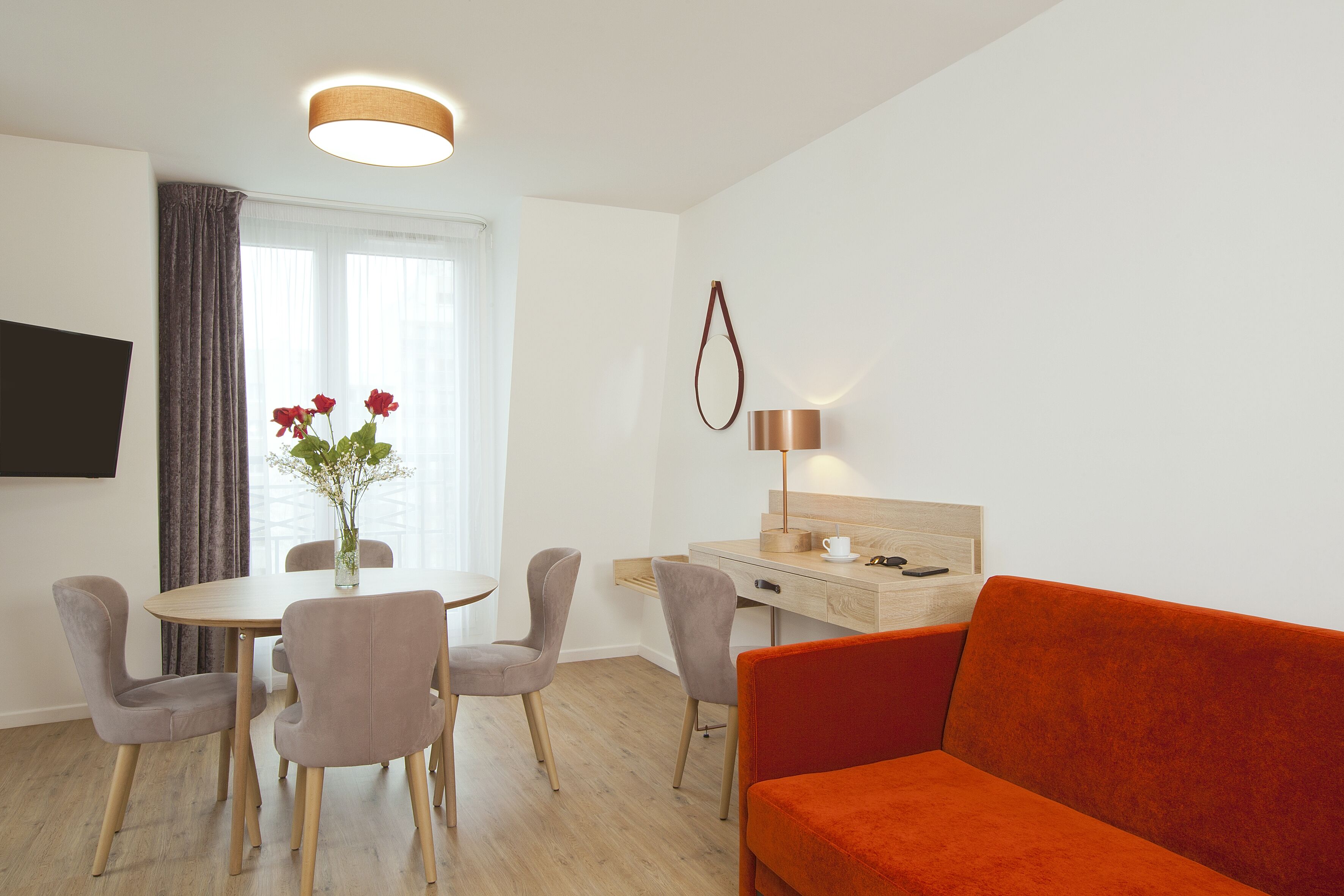 Una sala de estar moderna con una mesa de comedor de madera, cuatro sillas, un sofá, un televisor montado en la pared y un escritorio con una lámpara.