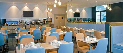 Un restaurante moderno con varias mesas y sillas, una zona de cocina y un gran ventanal que proporciona luz natural.