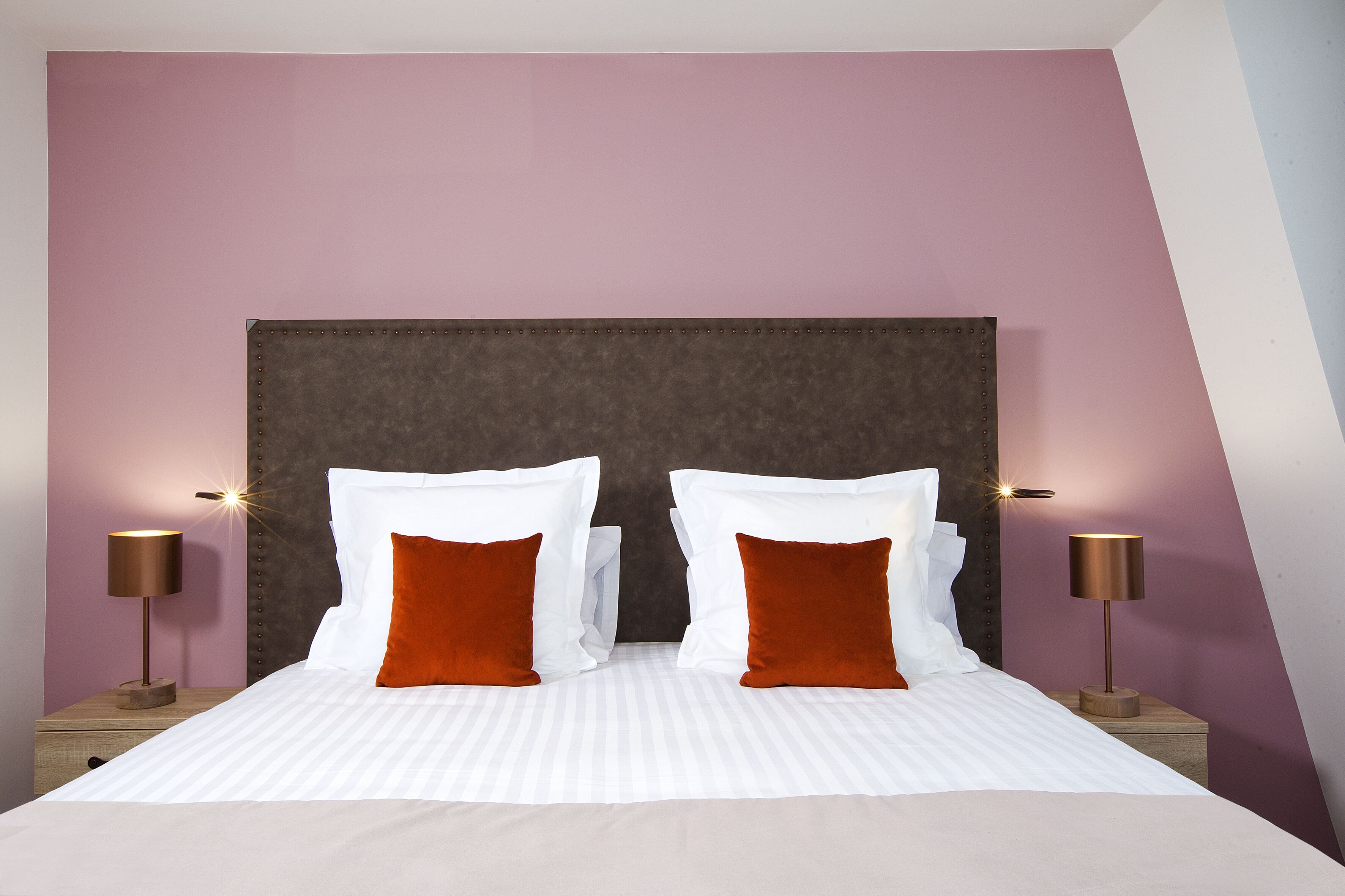 Una cama bien hecha con sábanas blancas y dos almohadas naranjas, flanqueada por dos mesitas de noche con lámparas, frente a una pared con cabecera oscura.