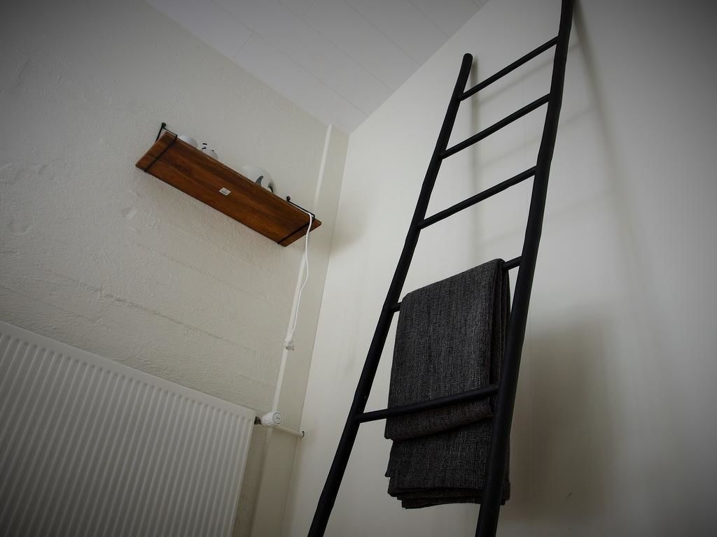 A room with a black metal ladder leaning against a white wall, a wooden shelf, and a radiator.