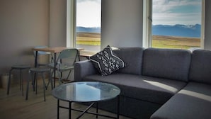 Un soggiorno moderno con un divano grigio, un tavolino in vetro e due poltrone. Ampie finestre offrono vista su un paesaggio pianeggiante con montagne in lontananza.