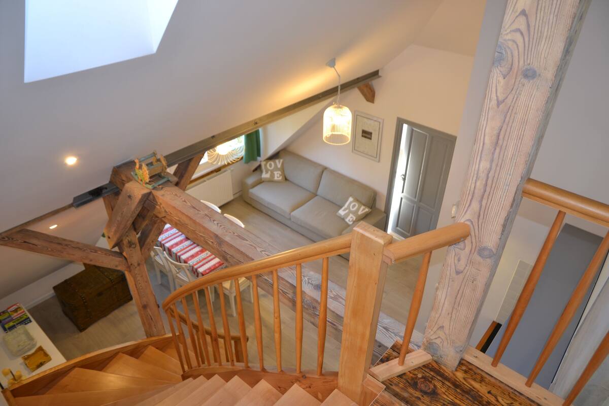 A wooden staircase leading to a loft bedroom with a sloped ceiling, a sofa with 'LOVE' pillows, and a hanging light fixture.