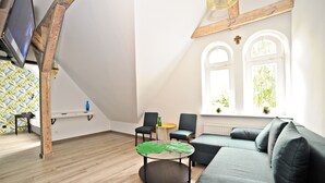 A spacious living room with a high ceiling, a grey sofa, two armchairs, a round coffee table with a leaf design, and a television mounted on the wall. - Grand Apartments - Loft (Sopot)