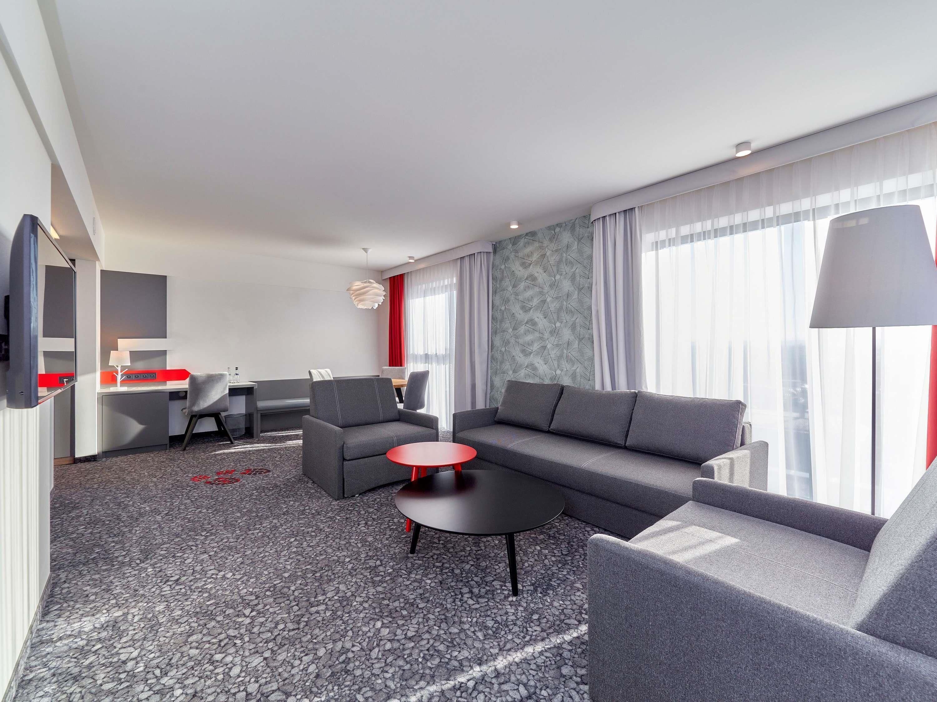 Ein modernes Hotelzimmer mit einer Couch, Sesseln, einem kleinen Tisch, einem Schreibtisch und einem Fernseher.