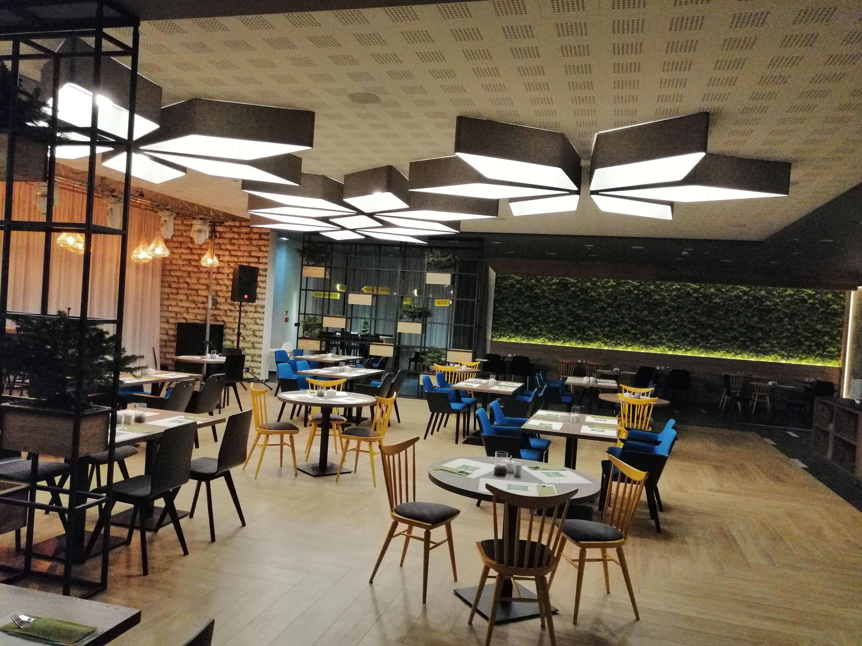 Interior modern d'un restaurant amb taules rodones, caires blaus i grocs, i una paret verda amb plantes.